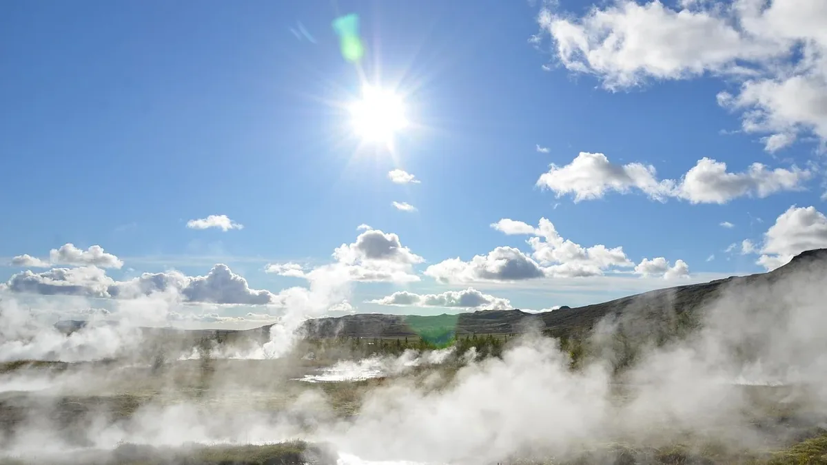 A mountainous landscape with steam rising from the ground, set against a blue sky with clouds and the sun shining brightly.