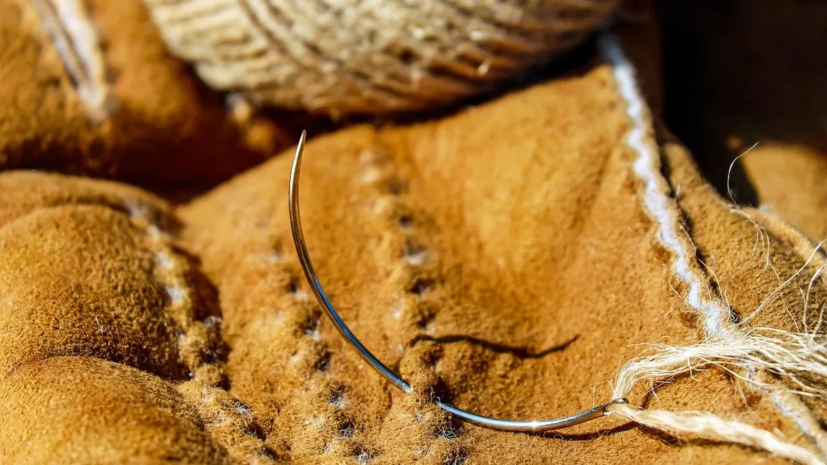 A close-up of a needle lying on a piece of tan fabric with visible stitching.