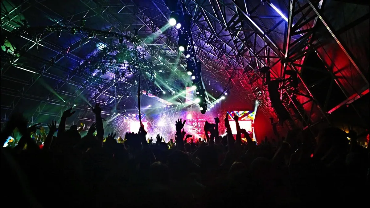 A crowd of people in a dark room with a stage featuring a DJ booth, surrounded by a metal structure and illuminated by various lights.