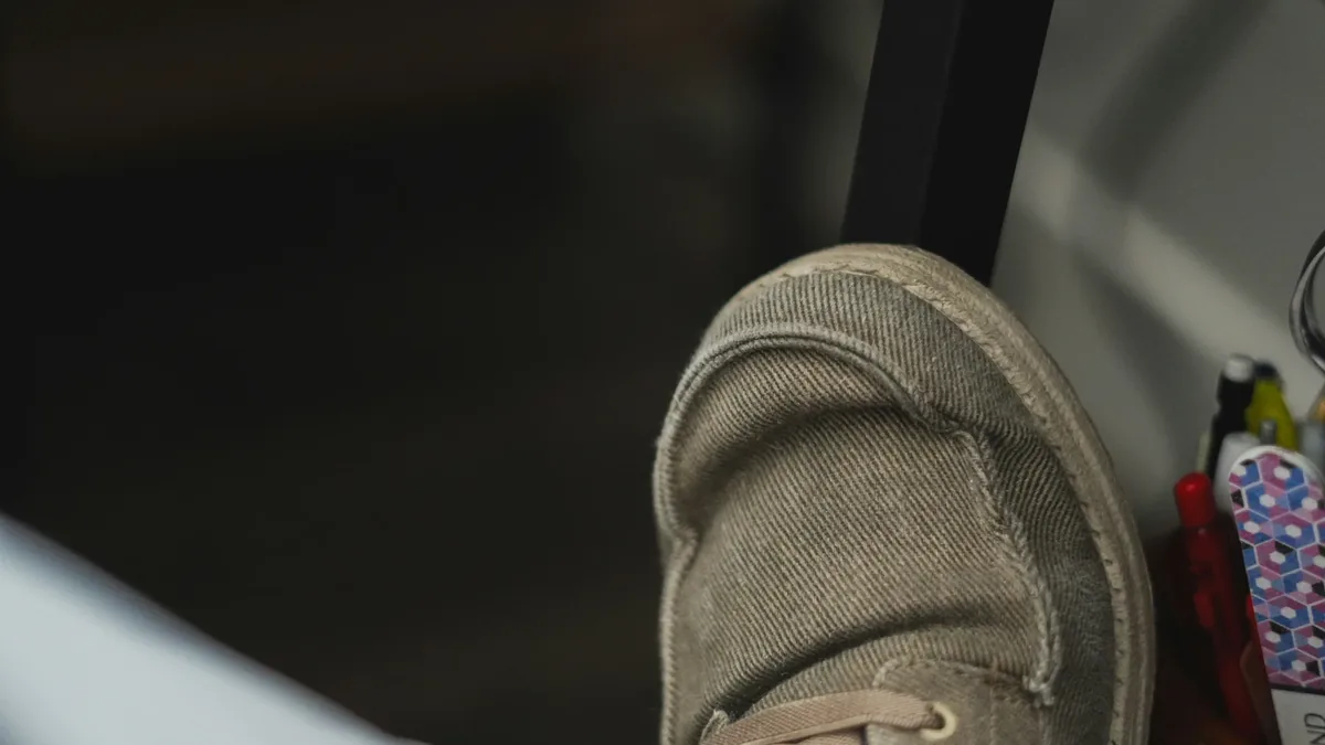 A close-up of a shoe, with a portion of a person's leg visible in the bottom left corner, and a shelf or table in the background.